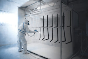 Worker wearing protective wear performing powder coating of metal details in a special industrial camera